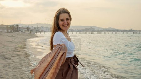 Téléchargez les photos : Portrait de joyeuse femme riante ludique court et tourne sur la plage de sable fin au coucher du soleil tout en regardant à la caméra - en image libre de droit