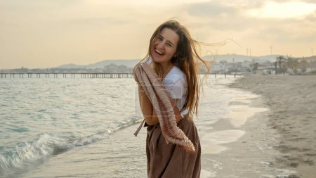 Photo for Happy young woman running on a sandy beach with a silk scarf flowing behind her. Concept of happiness, active lifestyle, travel, and vacation. - Royalty Free Image