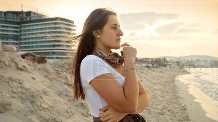 Téléchargez les photos : Une jeune femme réfléchie regardant vers la mer tout en tenant un foulard de soie coulant dans le vent sur une plage de sable au coucher du soleil - en image libre de droit