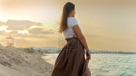 Téléchargez les photos : Jeune femme perdue dans la pensée, tenant une écharpe flottante en soie et regardant l'horizon océanique pendant le coucher du soleil sur la plage - en image libre de droit