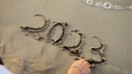 Téléchargez les photos : Gros plan de jeune femme écrivant 2023 nouvel an sur le sable humide à la plage de la mer. - en image libre de droit