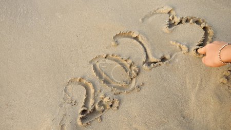 Téléchargez les photos : Gros plan de la main féminine en utilisant le doigt pour écrire 2023 année sur le sable humide à la plage de l'océan. - en image libre de droit