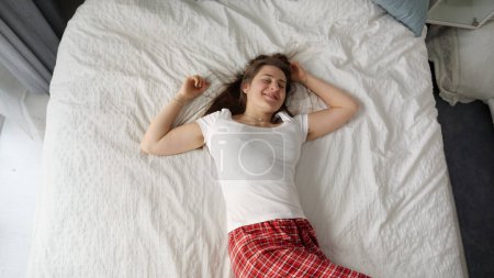 Foto de Mujer joven en pijama cae felizmente sobre una cama suave, expresión de alegría en su rostro. Concepto de relajación, ocio de fin de semana, y disfrutar de la comodidad del hogar - Imagen libre de derechos