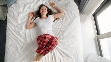 Photo for A woman in pajamas lying on a soft bed by a large window with a happy expression on her face. Concept of weekend leisure, relaxation, and cozy atmosphere at home - Royalty Free Image
