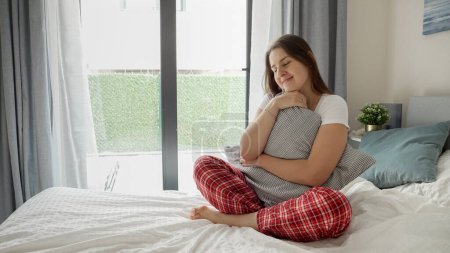 Téléchargez les photos : Jeune femme rêveuse en pyjama confortable assise sur le lit, embrassant un coussin et souriant à la pensée de son bien-aimé - en image libre de droit
