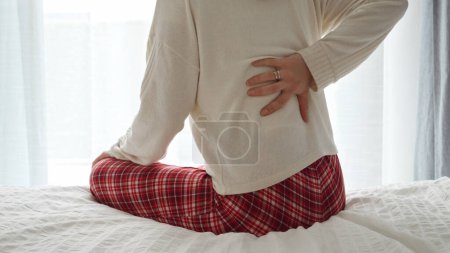 Téléchargez les photos : Jeune femme en pyjama assise sur le lit et tenant son bas du dos avec une douleur. Concept de problèmes de santé, d'inconfort et de gestion de la douleur - en image libre de droit