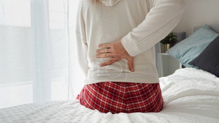 Photo for Young woman sitting on the edge of the bed in pajamas, rubbing her aching back. A concept of health problems, pain management, and self-care - Royalty Free Image