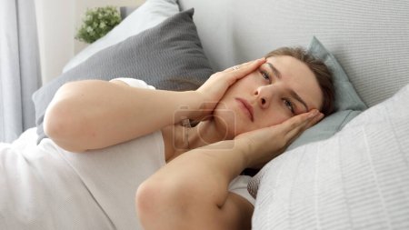 Téléchargez les photos : Portrait d'une jeune femme qui ressent une douleur à la tête en se tenant la main sur les tempes alors qu'elle est couchée au lit. - en image libre de droit