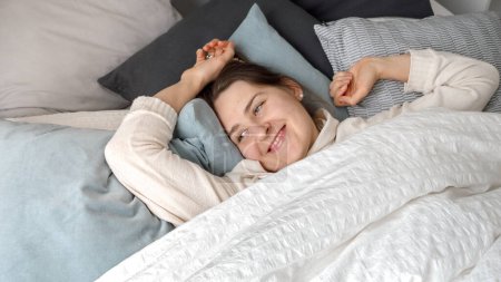 Photo for Portrait of smiling young woman opens eyes while lying and waking up in bed at morning. Concept of comfort, relaxation, healthy sleeping and good start of new day - Royalty Free Image