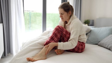 Foto de Mujer joven sintiendo calambre en la pierna acostada en la cama y masajeando los pies. Concepto de lesión, problemas de salud, dolor y trauma - Imagen libre de derechos