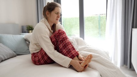 Téléchargez les photos : Jeune femme assise au lit et massant ses pieds douloureux à cause de crampes musculaires. - en image libre de droit