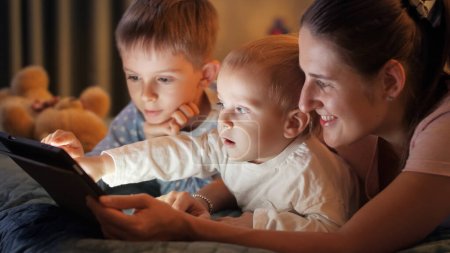 Foto de Madre joven acostada con dos niños en la cama jugando en la tableta por la noche. Familia pasando tiempo juntos, crianza, infancia feliz y entretenimiento - Imagen libre de derechos
