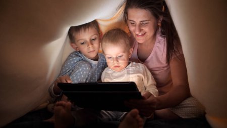 Téléchargez les photos : Famille heureuse en pyjama naviguant sur Internet et regardant la vidéo sur tablette dans le lit sous couverture. Famille ayant du temps ensemble, parentalité, enfance heureuse et divertissement - en image libre de droit