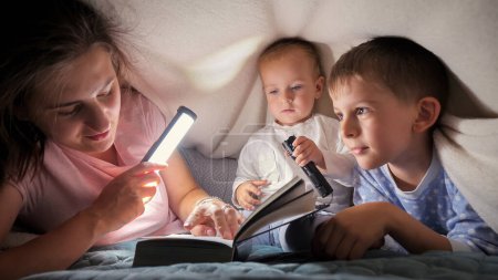 Foto de Joven madre cariñosa leyendo el libro de cuentos para sus dos hijos escondidos bajo una manta en la cama. Familia pasando tiempo juntos, crianza, infancia feliz y entretenimiento - Imagen libre de derechos