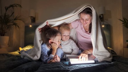 Photo for Two boys and mother in pajamas reading bedtime story book in bed at night with flashlights. Family having time together, parenting, happy childhood and entertainment - Royalty Free Image