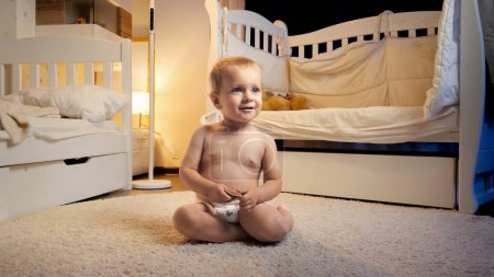 Photo for Cute baby boy in diapers sitting on carpet in children's room at night before going to sleep - Royalty Free Image