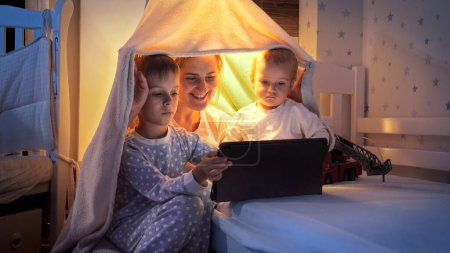 Foto de Joven madre viendo vídeo en la tableta con sus dos hijos en la cama por la noche. Familia pasando tiempo juntos, niños con gadgets, crianza, infancia feliz y entretenimiento. - Imagen libre de derechos