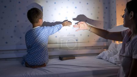Foto de Lindo chico con madre en pijama jugando con sombras en la pared desde la linterna. Familia pasando tiempo juntos, crianza, infancia feliz y entretenimiento - Imagen libre de derechos