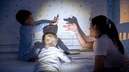 Téléchargez les photos : Des enfants joyeux avec leur mère jouant avec les ombres de la lampe de poche sur le mur la nuit. Famille ayant du temps ensemble, parentalité, enfance heureuse et divertissement - en image libre de droit