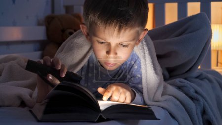 Foto de Retrato de niño con antorcha acostado en la cama y leyendo libro de cuentos. Educación infantil, desarrollo, secretismo, privacidad, lectura de libros - Imagen libre de derechos