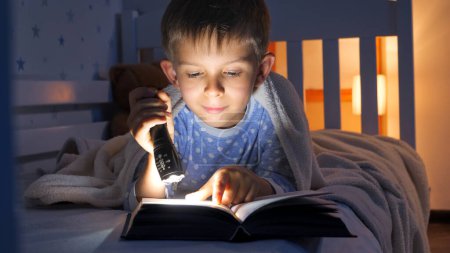 Foto de Niño en pijama acostado en la cama y leyendo un libro de cuentos con antorcha. Educación infantil, desarrollo, secretismo, privacidad, lectura de libros - Imagen libre de derechos
