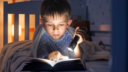 Foto de Retrato de un niño concentrado acostado en la cama y leyendo un libro con linterna. - Imagen libre de derechos