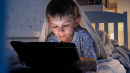 Téléchargez les photos : Portrait d'un petit garçon couché sous une couverture et utilisant un ordinateur tablette. Éducation des enfants, développement, enfants utilisant des gadgets secret, vie privée. - en image libre de droit