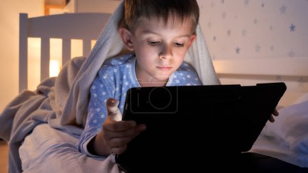 Photo for Portrait of cute boy in pajamas lying in bed and browsing internet on tablet computer. Children education, development, kids using gadgets secrecy, privacy. - Royalty Free Image