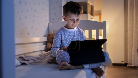 Téléchargez les photos : Petit garçon en pyjama assis avec tablette sur le lit et regarder la vidéo. Éducation des enfants, développement, enfants utilisant des gadgets secret, vie privée. - en image libre de droit