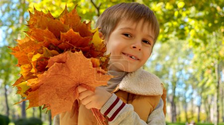 Foto de Retrato del alegre niño sonriente escondido detrás de un montón de hojas de arce amarillo en el parque de otoño. Niños al aire libre, niños felices e infancia, paisaje otoñal - Imagen libre de derechos