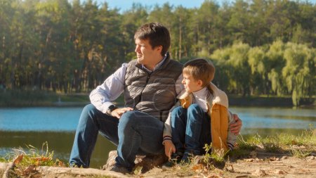 Foto de Joven padre cariñoso hablando con su hijo mientras está sentado en el lago en el bosque. Familia al aire libre, padres felices e infancia, paisaje otoñal, niños y padres relajándose al aire libre - Imagen libre de derechos