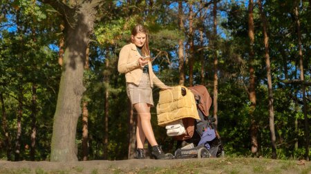 Foto de Mujer joven usando smartphone mientras camina con cochecito de bebé en el parque. Crianza, relajarse al aire libre, hermosa madre. - Imagen libre de derechos