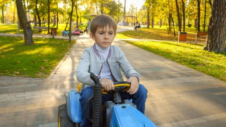 Téléchargez les photos : Portrait d'un petit garçon concentré conduisant un tracteur pour enfants au parc d'automne. Enfants jouant dans le parc, l'agriculture, le transport routier. - en image libre de droit