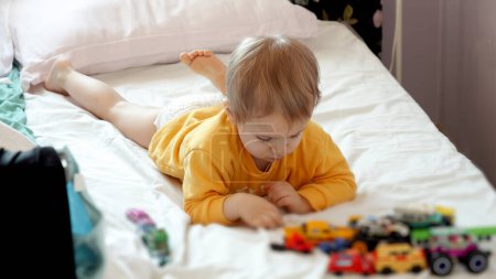 Foto de Lindo niño acostado en la cama y jugando con juguetes y coches de colores. Desarrollo infantil, educación y entretenimiento. - Imagen libre de derechos