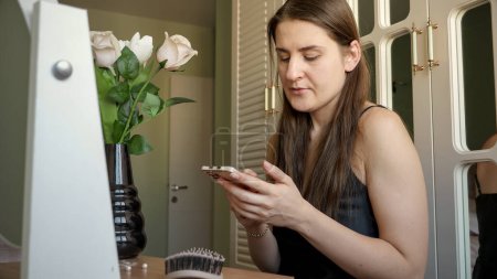 Photo for Young brunette woman sitting at makeup table in bedroom and using smartphone - Royalty Free Image