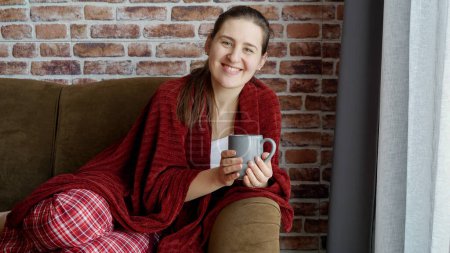 Téléchargez les photos : Heureuse femme souriante en pyjama reposant sur un canapé avec une tasse de thé. Femme au repos à la maison, mode de vie et détente - en image libre de droit