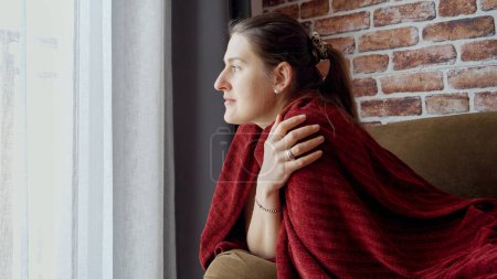 Téléchargez les photos : Jeune femme se sentant froide sur un canapé recouvert de carreaux rouges et regardant par la fenêtre. Femme au repos à la maison, mode de vie et détente - en image libre de droit