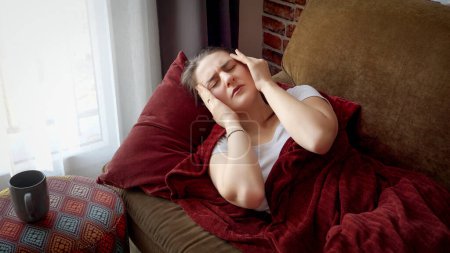 Foto de Mujer joven enferma que tiene la temperatura acostada en el sofá y tocándose la frente. Asistencia sanitaria, malestar de la mujer en casa, enfermedad y problemas de salud - Imagen libre de derechos