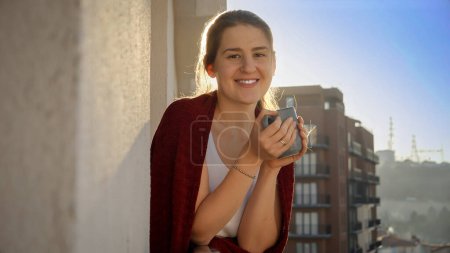 Photo for Happy smiling woman drinking tea on the balcony at morning. People relaxing, resting at home, beautiful cityscape - Royalty Free Image