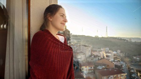 Téléchargez les photos : Femme brune souriante couvrant en plaid debout sur le balcon et regardant sur la terrasse. Les gens se relaxent, se reposent à la maison, beau paysage urbain - en image libre de droit