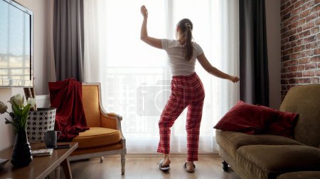 Téléchargez les photos : Photo drôle de femme heureuse portant un pyjama et dansant contre une grande fenêtre dans le salon. Les gens se détendre à la maison, avoir du plaisir, émotions positives et plaisir - en image libre de droit
