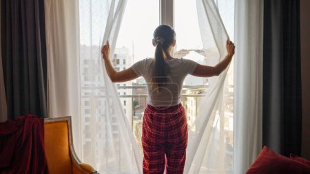 Photo for Rear view of woman in pajamas walking to big window in living room and opening curtains. People relaxing, resting at home, beautiful cityscape - Royalty Free Image