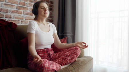 Photo for Young woman meditating and listening to relaxing music in headphones. Concept of relaxation, healthy lifestyle, resting at home and practicing yoga - Royalty Free Image