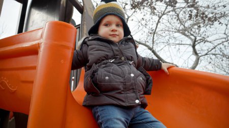 Téléchargez les photos : Mouvement lent du garçon tout-petit mignon glissant sur la glissière sur l'aire de jeux au parc public le jour d'automne froid - en image libre de droit