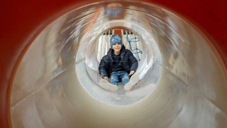 Foto de Pequeño niño deslizándose por el tobogán del tubo en el patio público. Niños felices, diversión al aire libre, ocio en el parque - Imagen libre de derechos