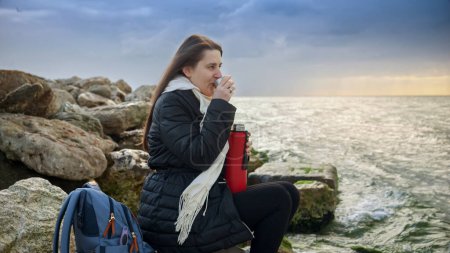 Foto de Mujer joven sintiendo frío sentada en los acantilados y mirando el frío mar del norte. Concepto de turismo, viajes, viajes, vacaciones y vacaciones en invierno - Imagen libre de derechos