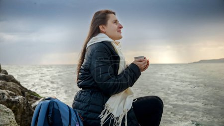 Foto de Mujer morena joven relajándose en el acantilado en el mar frío con taza de té caliente. - Imagen libre de derechos