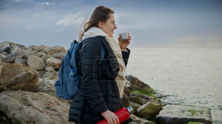 Téléchargez les photos : Femme aux cheveux longs, debout sur les rochers et regardant les vagues d'hiver tout en buvant du thé chaud. Un excellent ajout aux voyages ou aux projets sur le thème des vacances - en image libre de droit