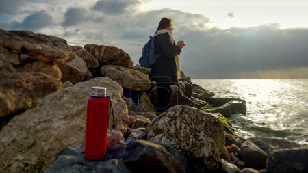 Foto de Joven excursionista de pie en los acantilados en la costa del mar y beber té caliente mientras mira la puesta de sol sobre el océano - Imagen libre de derechos