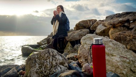 Téléchargez les photos : Jeune femme se détendre sur la côte rocheuse au coucher du soleil et boire du thé chaud par temps froid et venteux. Concept de tourisme, voyage, voyage, vacances et vacances en hiver - en image libre de droit